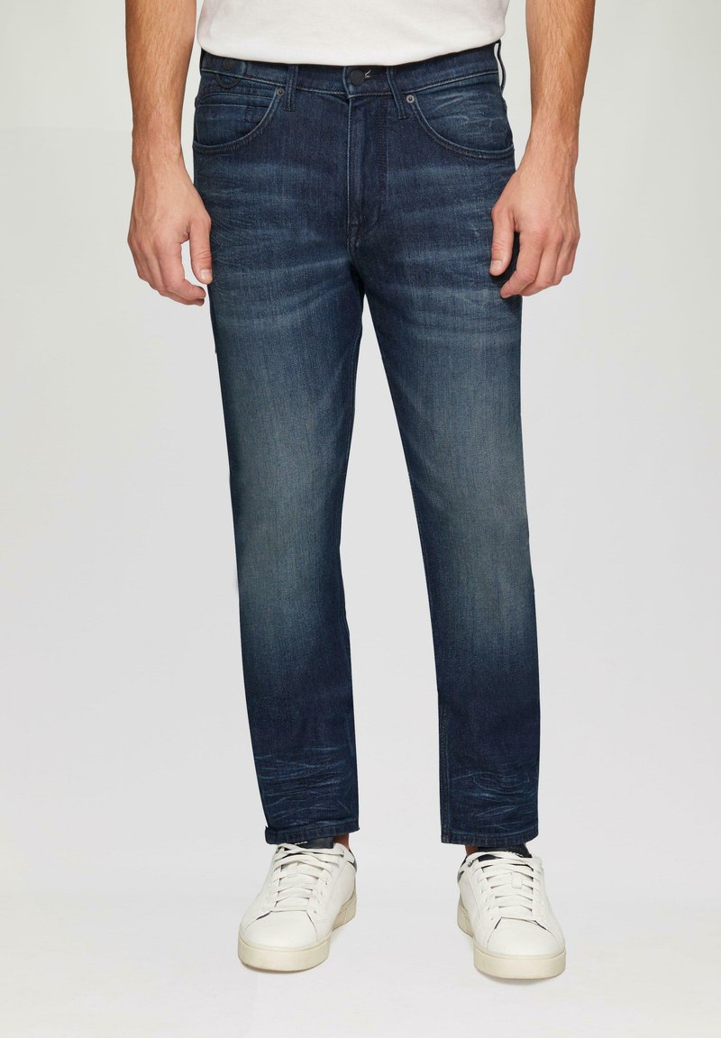Jean en denim bleu foncé à coupe slim, avec cinq poches, un léger délavage et une fermeture à bouton, portés avec des baskets blanches.