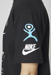 Černé bavlněné tričko s modrou grafikou na rameni a bílým logem Nike. Krátké rukávy a volný střih.