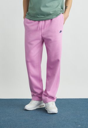 Mężczyzna w jasnofioletowych spodniach dresowych Nike, zielonej koszulce i białych tenisówkach stoi na niebieskim dywanie z rękami w kieszeniach.