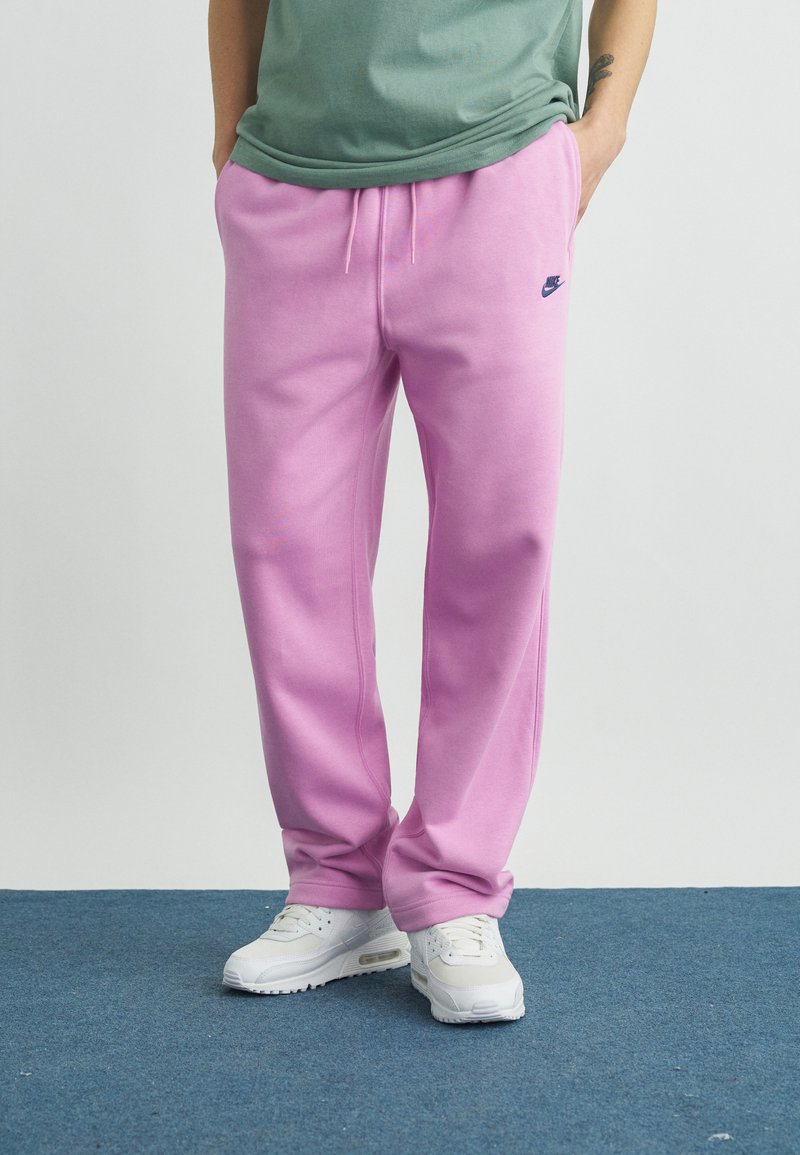 Muž stojaci na modrom koberci, oblečený v svetlofialových teplákoch Nike, zelenej tričku a bielych teniskách, s rukami v vreckách.