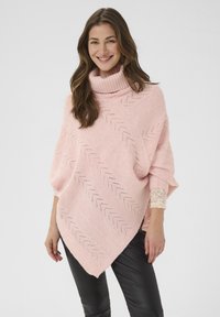 Ženska z dolgimi rjavimi lasmi se smeji, oblečena v roza pleteni pončo s turtleneckom in chevron vzorcem, ki ga nosi čez bluzo z čipkastimi rokavi in črne hlače.