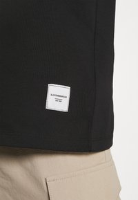 Sort kortærmet t-shirt med et firkantet hvidt mærke, der har "Lindbergh" og "Est. 1927" broderet på den nedre søm, set fra siden.
