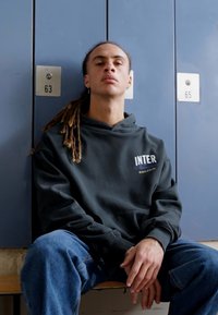 Sweatshirt preto com capuz e o texto "INTER", combinado com jeans de ganga azuis. Indivíduo sentado em frente a armários cinzentos numerados 63 e 65.