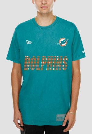 Mann trägt ein türkisfarbenes Miami Dolphins T-Shirt mit Teamlogo auf der Brust und gestreiftem "DOLPHINS" Text auf der Vorderseite.