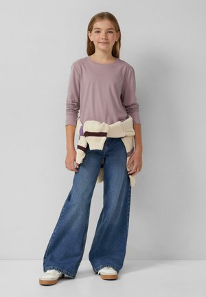 Lange mouw lavendelkleurige top gecombineerd met wijde blauwe jeans. Een gebreide, crèmekleurige trui rondom de taille geknoopt, met paarse en bruine accenten.