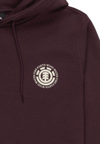 Sudadera color marrón con capucha que presenta un logo circular crema en el pecho, con texto estilizado alrededor de un diseño central de árbol. Textura de tela suave.