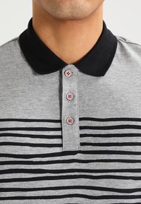 Šedé polo tričko s černým límečkem a vodorovnými černými pruhy. Pět knoflíků vpředu, s bledými knoflíkovými dírkami.