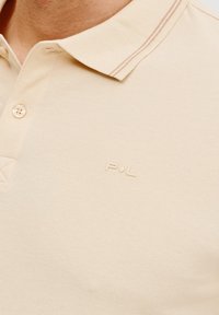 Beżowa koszulka polo z teksturowanej tkaniny, z kołnierzykiem na guziki i cienkimi paskami. Dyskretne logo P/VL na piersi.