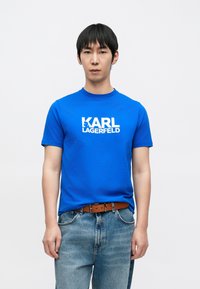 Zils kokvilnas T-krekls ar apaļu apkakli un īsām piedurknēm, priekšpusē izcelts balts "KARL LAGERFELD" logotips. Valdāts ar džinsa biksēm.