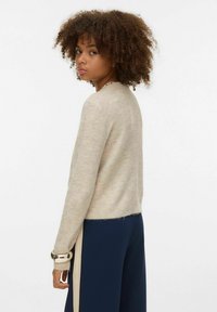 Béžový pletený svetr s kulatým výstřihem, mírně zkrácené délky a třepícím se lemem. Nošen spolu s tmavě modrými kalhotami s bočním proužkem.