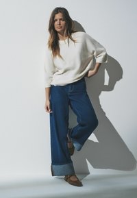 Bela pletena majica z širokimi rokavi, v kombinaciji s temno modrimi širokimi hlačami z zavihanimi robovi in rjavimi balerinkami. Preprost, čist dizajn.