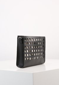 Černá kožená taška s vyřezávaným vzorem, plochý obdélníkový tvar, hladká textura a boční připevnění popruhu. Minimalistický design.