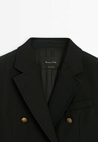 Musta bleiseri, jossa on jämäkkä kaulus, kaksi kultanappia ja tasku. Sisällä oleva merkki, jossa lukee "Massimo Dutti", valmistettu Marokossa. Sileä rakenne.