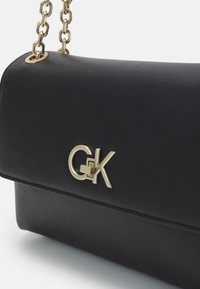 Musta nahkainen käsilaukku, jossa on kultaiset yksityiskohdat, ketjuolkahihna ja etupuolella näkyvä suuri kultainen "G&K"-logo. Sileä pinta.