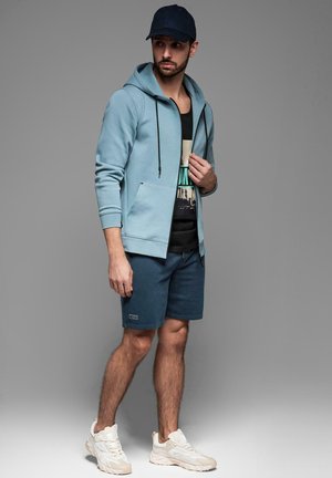 Jasnoniebieska bluza z kapturem na zamek z przednią kieszenią, czarny top na ramiączkach pod spodem, ciemnoniebieskie szorty oraz kremowe sneakersy, uzupełnione granatową czapką.