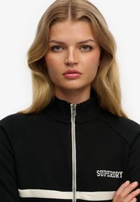 Czarna bluza rozpinana na zamek z białym paskiem i logo, z wysokim kołnierzem oraz prążkowaną teksturą na ramionach i mankietach.