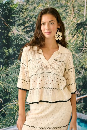 Mladá žena s dlhými hnedými vlasmi, ktorá má na sebe krémový pletený top a sukňu s čiernym lemom, veľký krémový kvetinový náušník, na pozadí zelených listov.