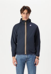Navy windbreaker jacket with a front zipper, yellow and red stripe accents, and a drawstring hood. Worn over light blue jeans.