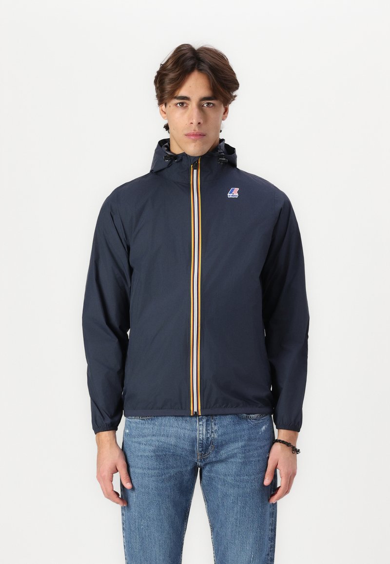 Navy windbreaker jacket with a front zipper, yellow and red stripe accents, and a drawstring hood. Worn over light blue jeans.