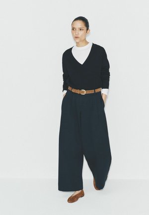 Frau steht und trägt schwarze Hosen mit weitem Bein, schwarzen V-Ausschnitt-Pullover über einem weißen Oberteil, braunen Gürtel und braune Ballerinas.