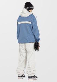 Veste extérieure bleu et blanc avec capuche, dotée de poignets élastiques, associée à un pantalon blanc ample et des baskets sombres.