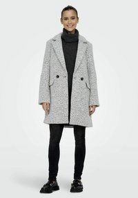 Manteau gris texturé à double boutonnage avec larges revers, poches latérales et deux boutons noirs, porté par-dessus un pull en tricot sombre et un jean skinny noir.
