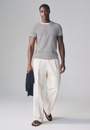 Mand iført hvidt stribet t-shirt og løse hvide bukser, der holder en mørk jakke i den ene hånd, går fremad på en lys grå baggrund.