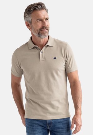 Homme d'âge moyen aux cheveux gris et à la barbe, portant un polo beige et un jean bleu, regardant vers la droite avec un léger sourire.