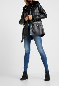 Chaqueta de cuero negra con capucha forrada de piel, con bolsillos con cremallera y solapa, combinada con vaqueros azules desgastados y botines negros.