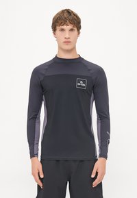 Rash guard z długim rękawem w ciemnoszarym kolorze z czarnym panelem na klatce piersiowej, białym logo i jasnoszarymi akcentami po bokach, wykonany z elastycznego materiału odprowadzającego wilgoć.