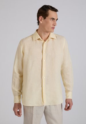Mężczyzna ubrany w jasnożółtą lnianą koszulę na guziki z długim rękawem oraz jasnobeżowe spodnie, patrzący w prawo na jednolitym tle.