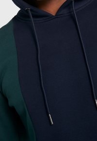 Felpa con cappuccio di colore blu navy con maniche verdi. Presenta lacci con punte in metallo e un tessuto morbido e texturizzato.