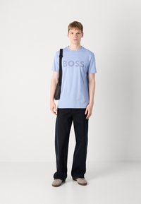 T-shirt en coton bleu clair avec le logo "BOSS" en bleu plus foncé, associé à un jean foncé. Les accessoires comprennent un sac à bandoulière noir et des chaussures claires.