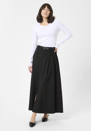 Gonna nera lunga fino ai piedi con vita alta, pieghe e uno spacco laterale. Abbinata a una camicia fitted bianca a maniche lunghe e scarpe nere a punta.