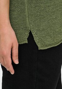 Alyvuogių spalvos megztinis su tekstūruota rašte, su šoninėmis plyšėmis ir apvaliais kraštais, derinamas su juodomis kelnėmis. Rankos matoma dydžio palyginimui.