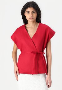 Top rosso avvolgente in lino con scollatura a V, maniche ampie e una cintura in tessuto in vita. Gonna bianca a trama completa l'outfit.
