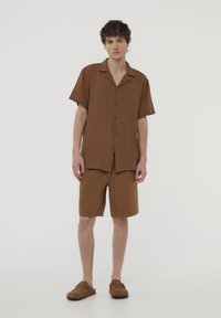 Camicia marrone a maniche corte con bottoni, pantaloni corti marroni abbinati e scarpe slip-on marroni. Tessuto morbido, vestibilità comoda e design semplice.