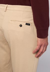 Beige Chinos aus Baumwoll-Twill, mit einer Gesäßtasche, die einen schwarzen Knopf und ein schwarzes Etikett mit dem Schriftzug "Trussardi" hat. Glatte Textur.