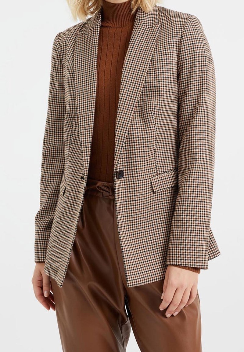 Femme portant un blazer marron à carreaux sur un pull à col roulé marron côtelé et un pantalon en cuir marron taille haute, debout devant un fond blanc.