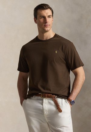 Mand iført mørkebrun kortærmet skjorte, hvide bukser med flettet brunt bælte og et armbåndsur, stående mod en neutral baggrund.