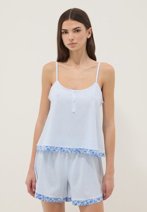 Giovane donna che indossa un top senza maniche a righe azzurro chiaro e bianco e pantaloncini abbinati con rifinitura floreale, in piedi davanti a uno sfondo semplice.