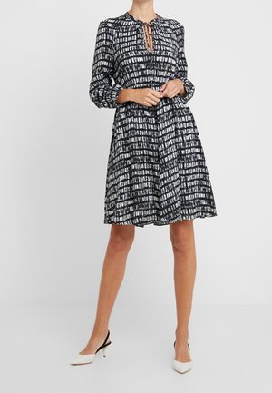 Kvinde iført knælange sort-hvide mønstrede kjole med bindebånd ved halsen og hvide slingback-hæle, stående mod en ensfarvet baggrund.