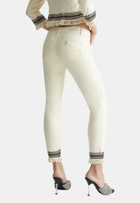 Jeans ajustados color crema con acentos decorativos de flecos en blanco y negro en los dobladillos. Bolsillos traseros visibles. Combinados con mules negros de tacón.
