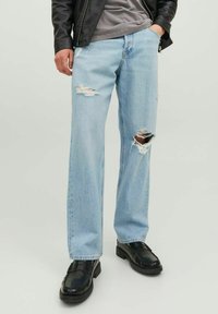 Jeans de mezclilla desgastados en azul claro con piernas anchas, con detalles rasgados. Llevados con zapatos de cuero negros y una camiseta gris debajo de una chaqueta negra.
