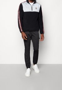 Černý fleecový pulovr s šedou horní částí, čtvrtinovým zipem, červeným bočním pruhem a logem. V kombinaci s černými tapered kalhotami a bílými teniskami.