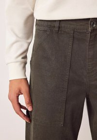 Dunkelolivgrüne Hose aus strukturiertem Stoff, mit seitlicher Tasche und Knopfverschluss. Die sichtbare Hand hält den Stoff.