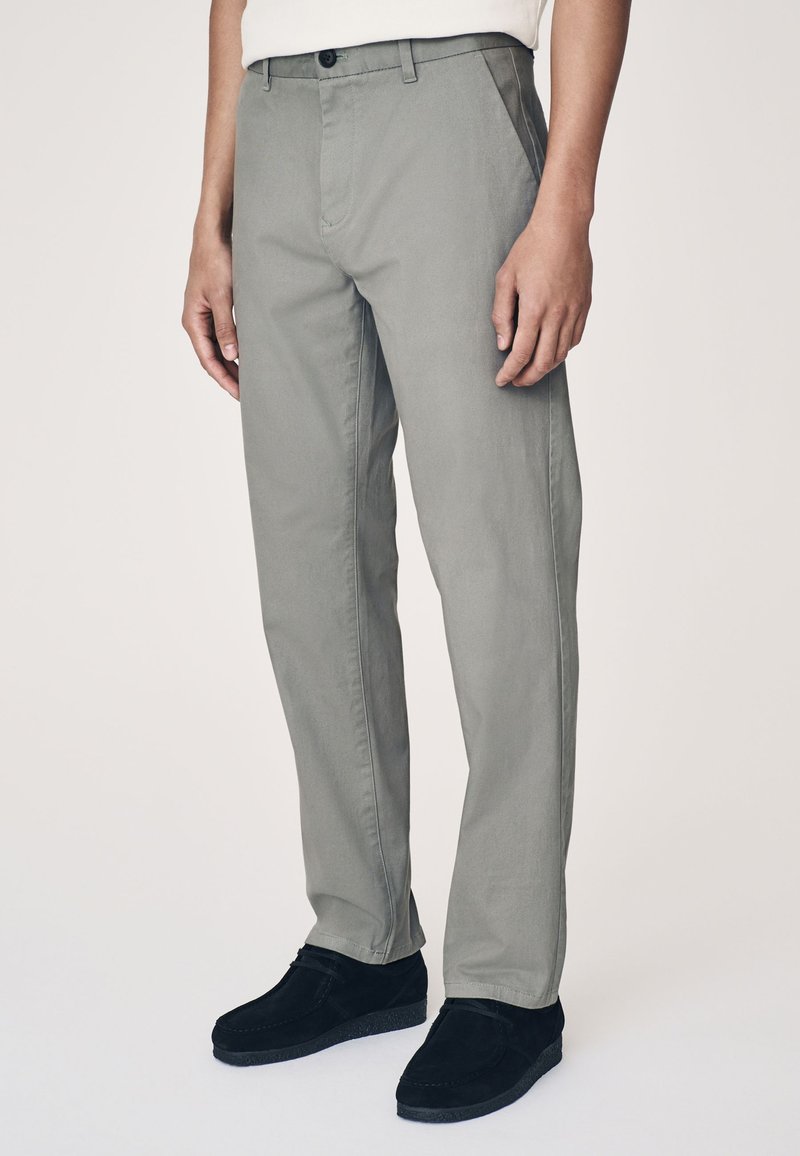 Pantaloni grigi chiari su misura realizzati in tessuto morbido, con design a gamba dritta, tasche frontali e chiusura con bottone. Indossati con scarpe nere.