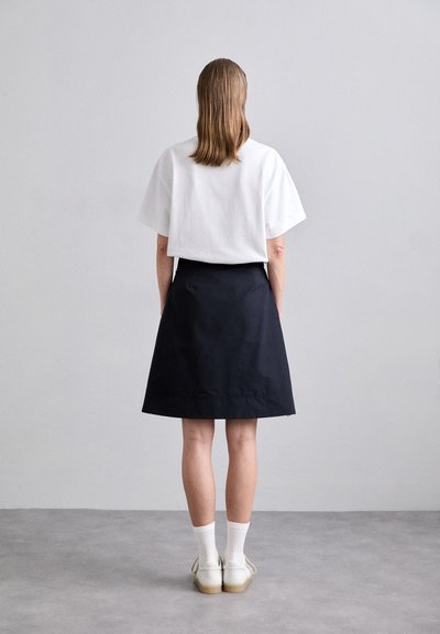 Weißes kurzärmeliges Baumwolloberteil, kombiniert mit einem schwarzen knielangen Rock. Model trägt weiße gerippte Socken und beigen Schuhe. Einfaches Design, einfarbige Farben.