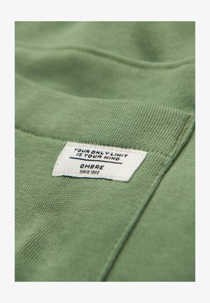 Grüner Strickstoff mit einem weißen angenähten Etikett, auf dem "YOUR ONLY LIMIT IS YOUR MIND" und "OMBRE since 1985" an einem gefalteten Rand steht.
