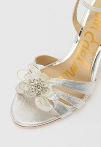 Silver high-heeled sandal featuring metallic straps, a textured finish, and a floral embellishment with transparent petals and silver accents.